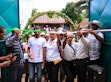 A group cuts a pink ribbon at the entrance to the Mirohy Center in Masindrano, framed by open turquoise gates and a leafy arch, while other participants look on.