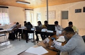 Workshop participants sit around tables arranged in a U-shape, reading papers and working on laptops in a classroom with wooden ceilings and curtained windows.