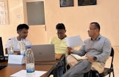 Three participants sit around a table during a discussion. One holds up a sheet of paper while the others look on. A laptop and notebooks lie on the table.
