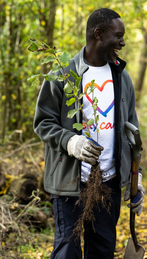 Volunteer holds a young tree and spade, smiling in a wooded area during a Sky Cares community project.