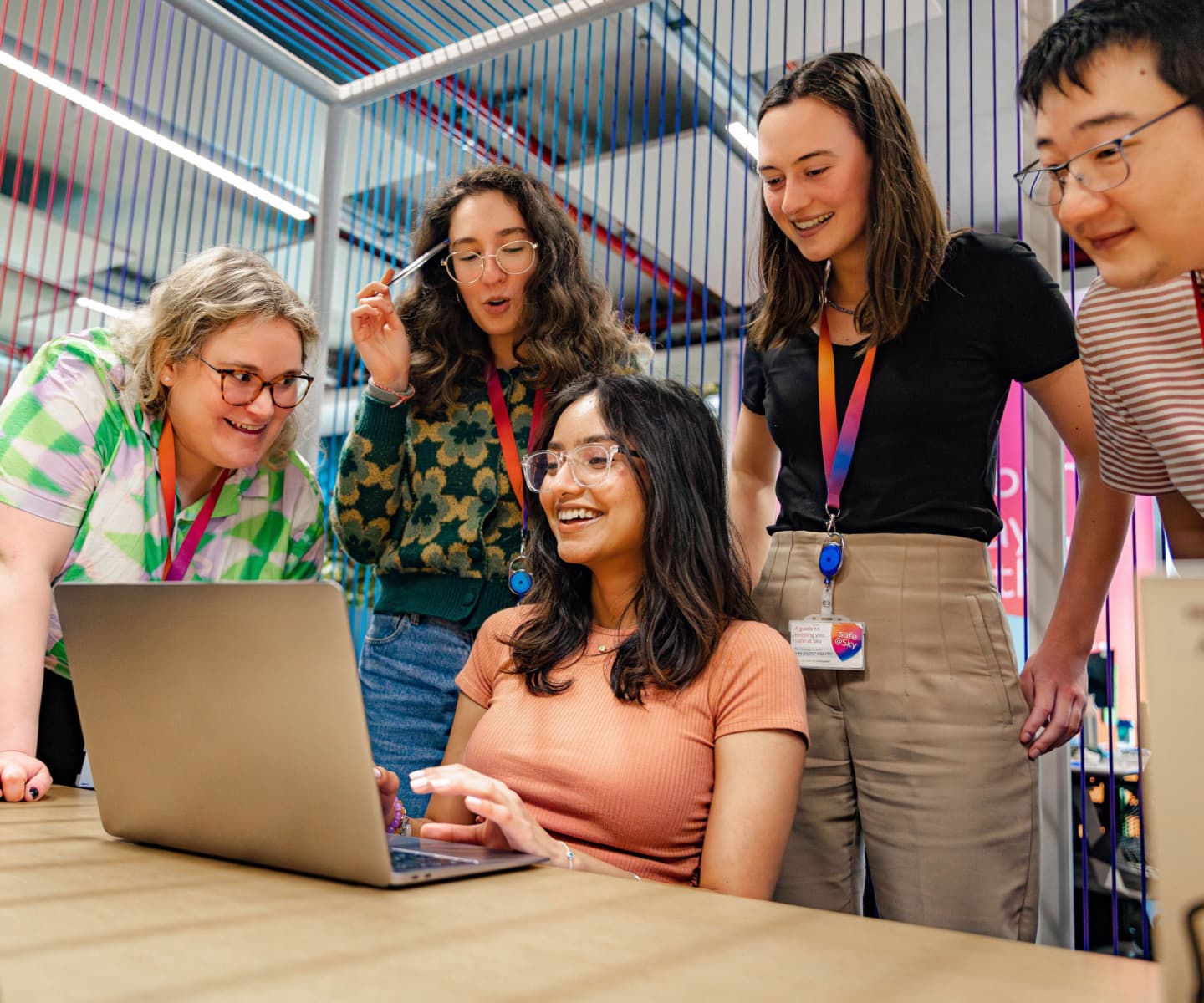 Five team members gather around a laptop, smiling and engaged in a creative, colorful workspace.