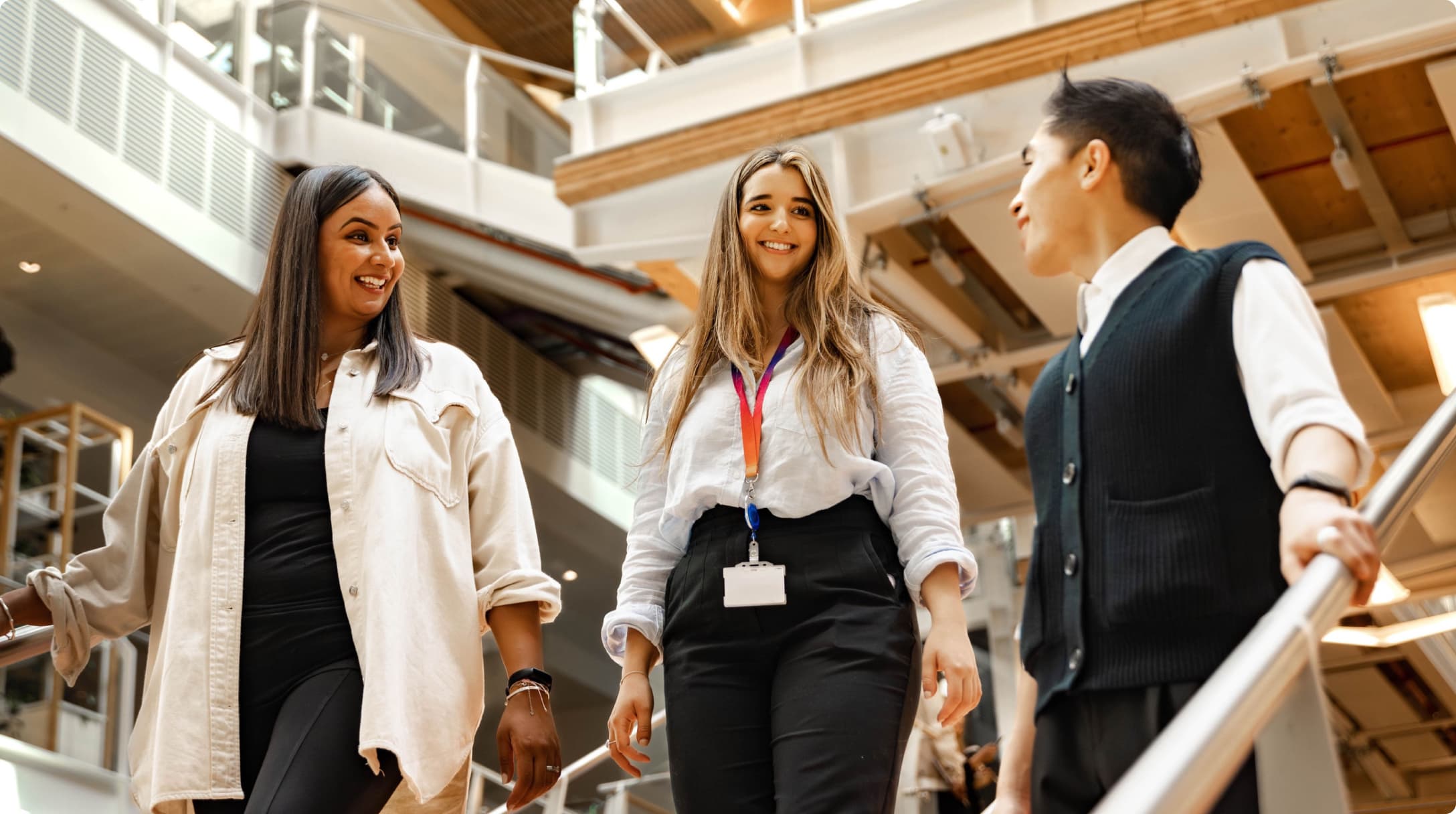 Three colleagues walk down a staircase, chatting and smiling in a modern office atrium with glass railings.