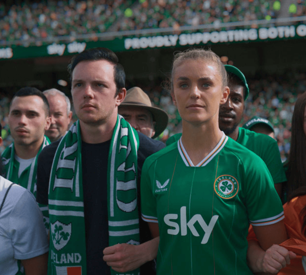 Football fans and players in green Ireland kits at a packed stadium, proudly supported by Sky.