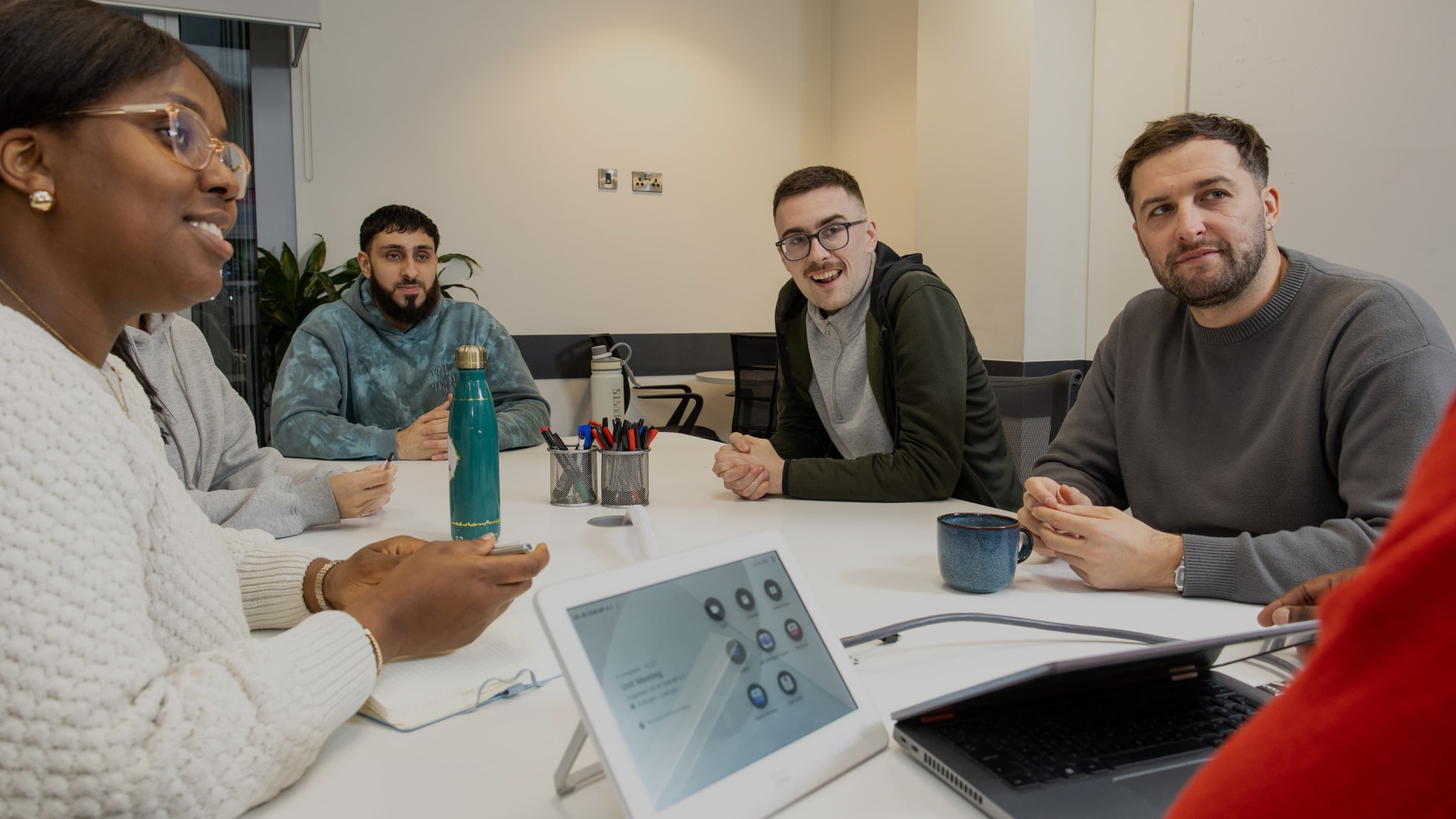 Team gathers in a bright meeting room, engaged in discussion around a white table with laptops and water bottles