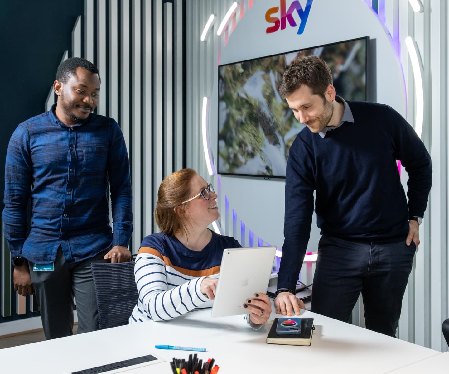 Three colleagues smile while collaborating on a tablet, standing in front of a Sky logo in a sleek workspace