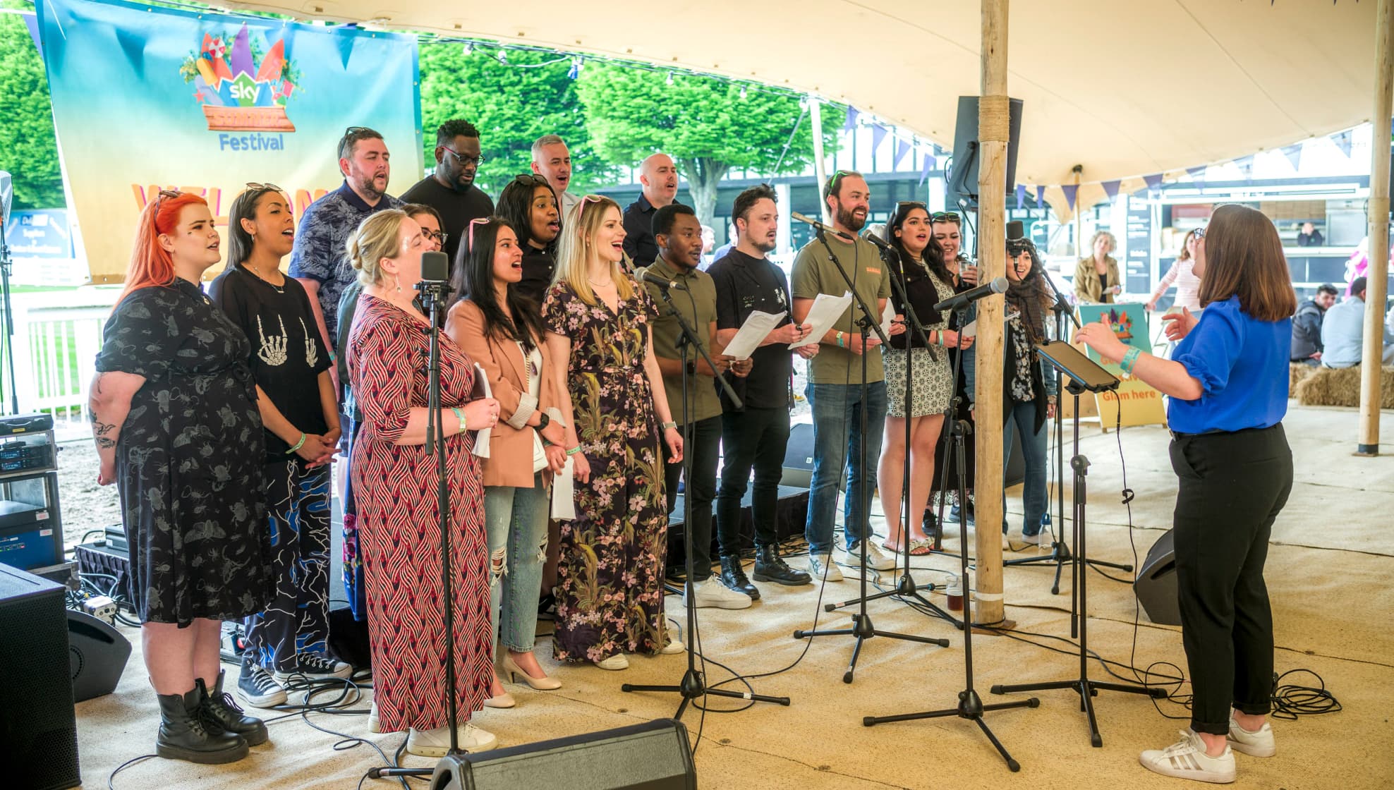 Choir group sings under a tent at a Sky summer event, led by a conductor, with microphones and instruments.