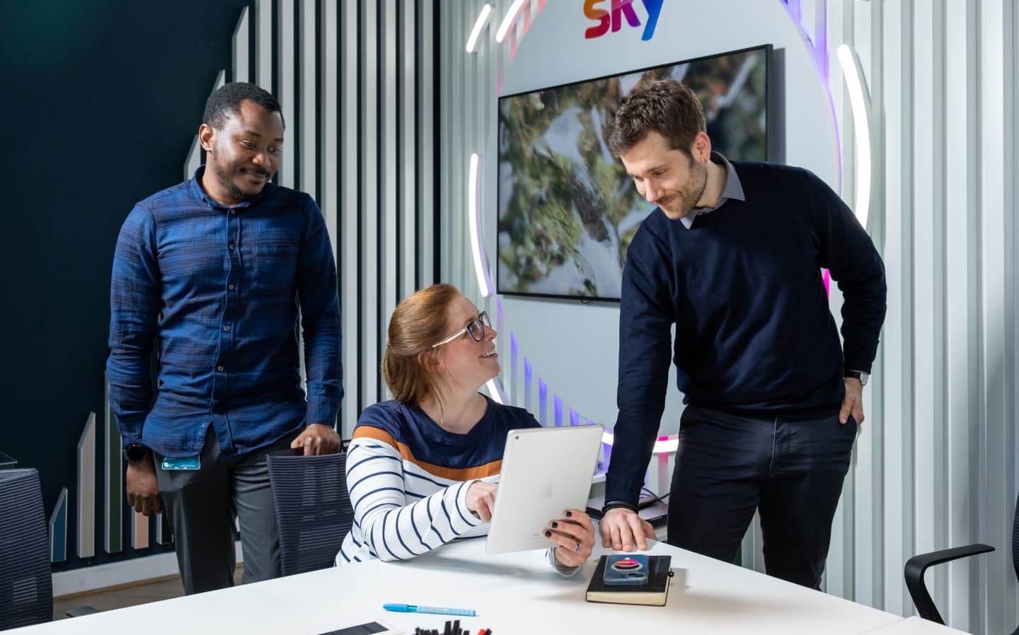 Three colleagues smile while collaborating on a tablet, standing in front of a Sky logo in a sleek workspace.