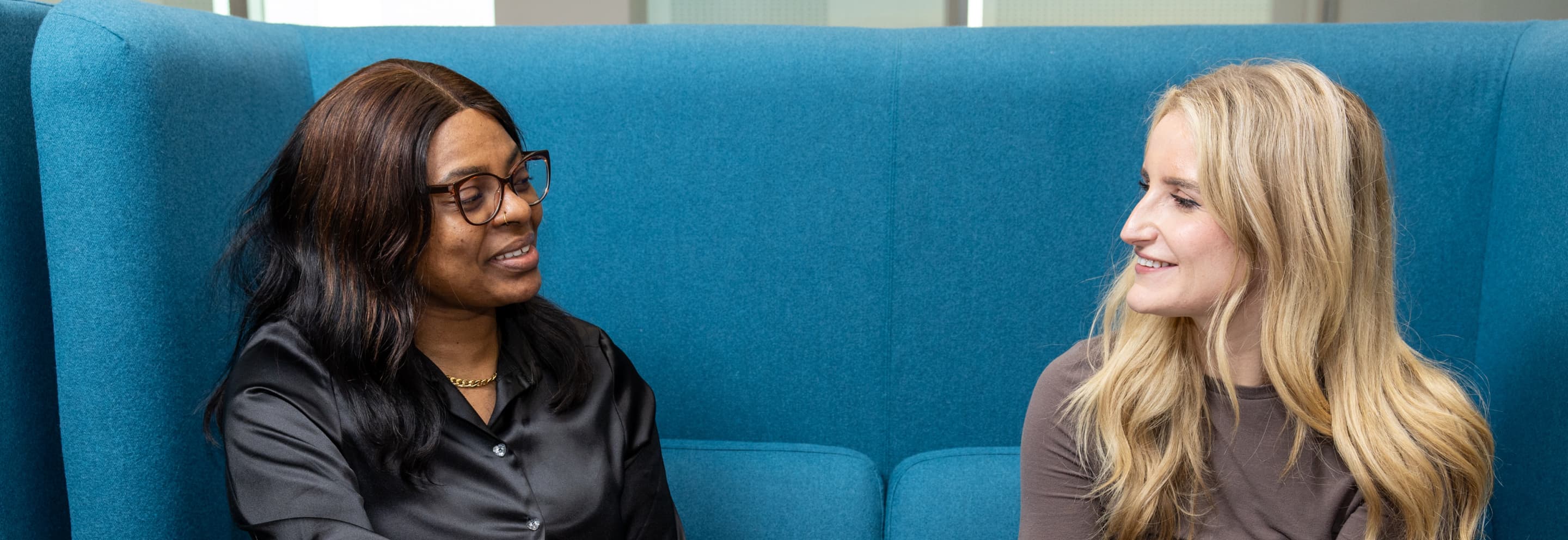 Two colleagues seated on a blue sofa, smiling and chatting in a relaxed, modern office space.