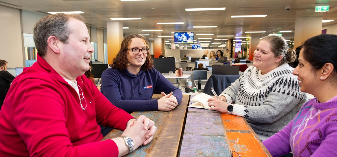 Four colleagues chat at a rustic wooden table in a modern office with laptops and notebooks around them.