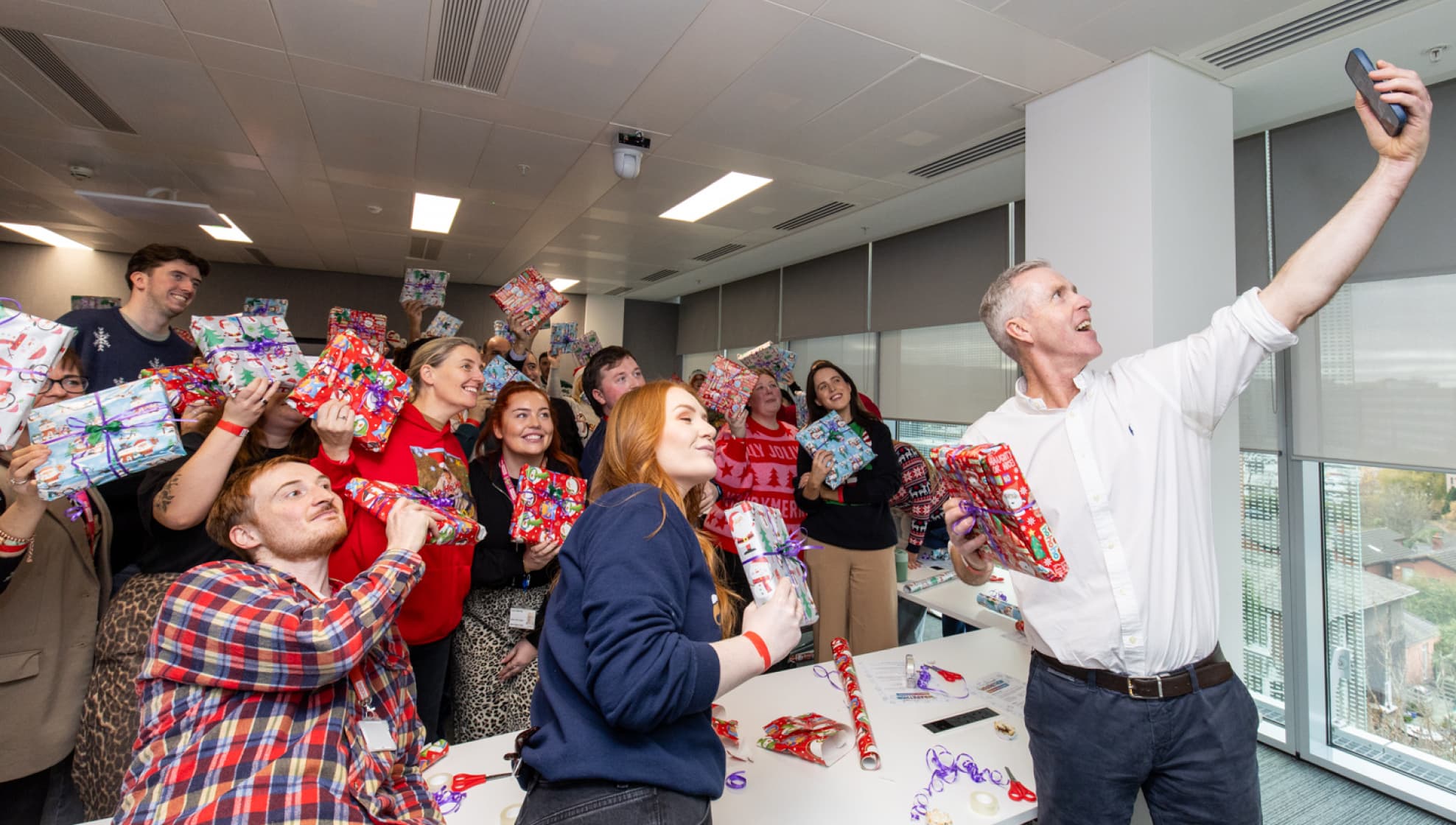 Team members in festive jumpers pose with wrapped gifts, smiling as a selfie is taken during a holiday event.
