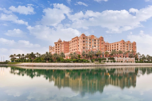 Luxury hotel with pink architecture reflecting in calm water under a partly cloudy sky.