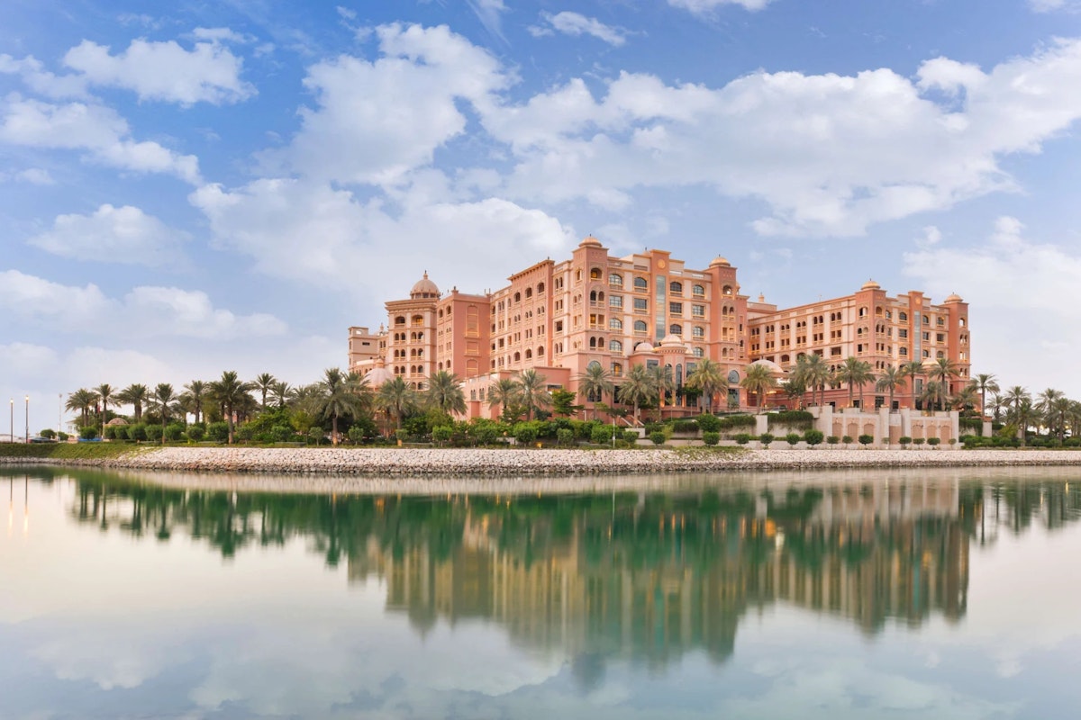 Luxury hotel with pink architecture reflecting in calm water under a partly cloudy sky.