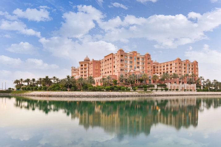 Luxury hotel with pink architecture reflecting in calm water under a partly cloudy sky.