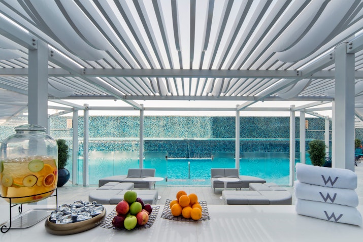 W Doha Poolside seating area with fresh fruits, towels, and drinks under a shaded pergola.