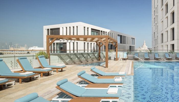 Alwadi Doha MGallery Hotel Collection Rooftop pool with lounge chairs, a wooden pergola, and a modern building in the background.