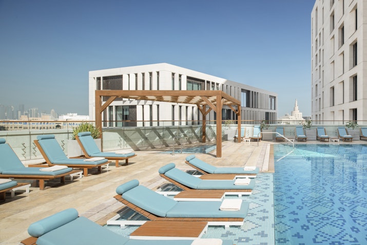 Rooftop pool with lounge chairs, a wooden pergola, and a modern building in the background.