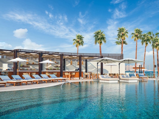 Resort pool surrounded by lounge chairs, palm trees, and shaded cabanas under a blue sky