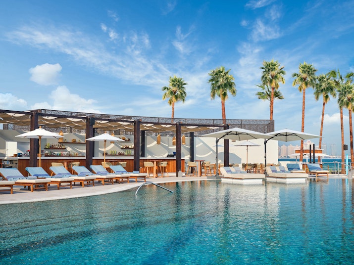 Resort pool surrounded by lounge chairs, palm trees, and shaded cabanas under a blue sky