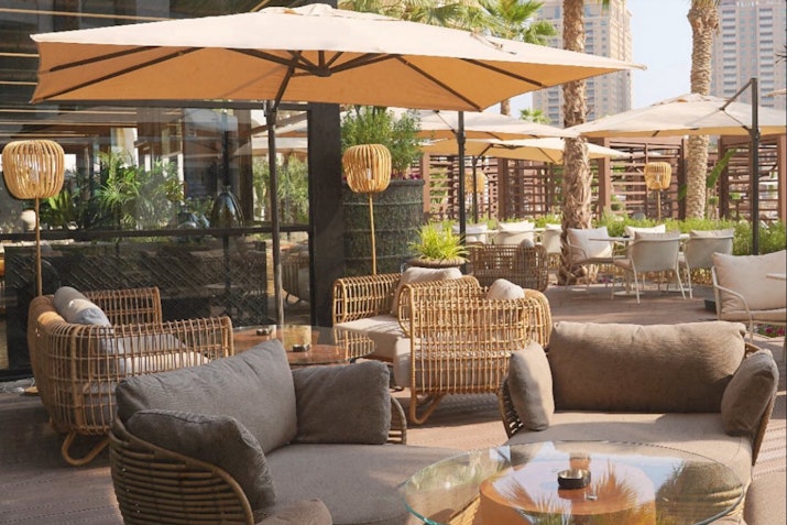Outdoor seating area with rattan furniture, large umbrellas, and palm trees in the background.