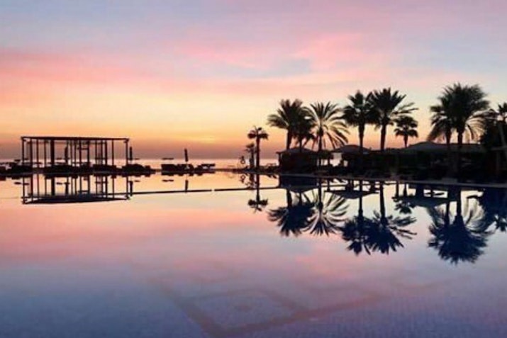 Infinity Rooftop Lounge at Alwadi Hotel Doha - MGallery Serene sunset over a reflective pool with palm trees at a resort.