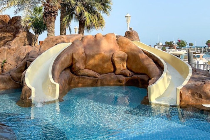 Water slides at a resort pool surrounded by palm trees and a scenic view.