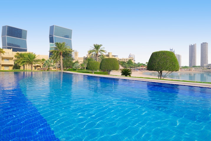 Outdoor pool with clear blue water, palm trees, and modern buildings in the background