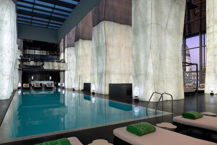 Indoor swimming pool with marble walls, lounge chairs, and modern lighting.