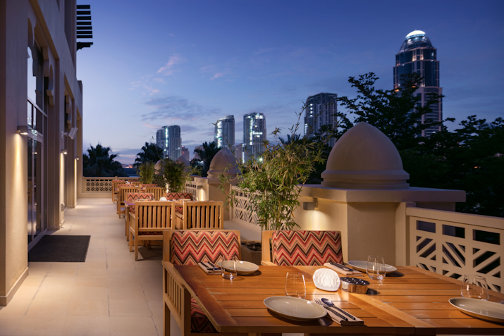 Outdoor dining area with wooden tables, cozy seating, and a view of the city skyline at dusk.
