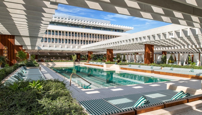 The Ned Doha Modern pool area with lounge chairs and shaded pergolas, surrounded by lush greenery and a building.