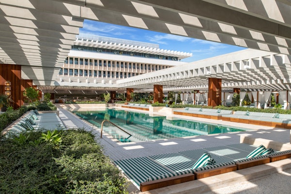 Modern pool area with lounge chairs and shaded pergolas, surrounded by lush greenery and a building.