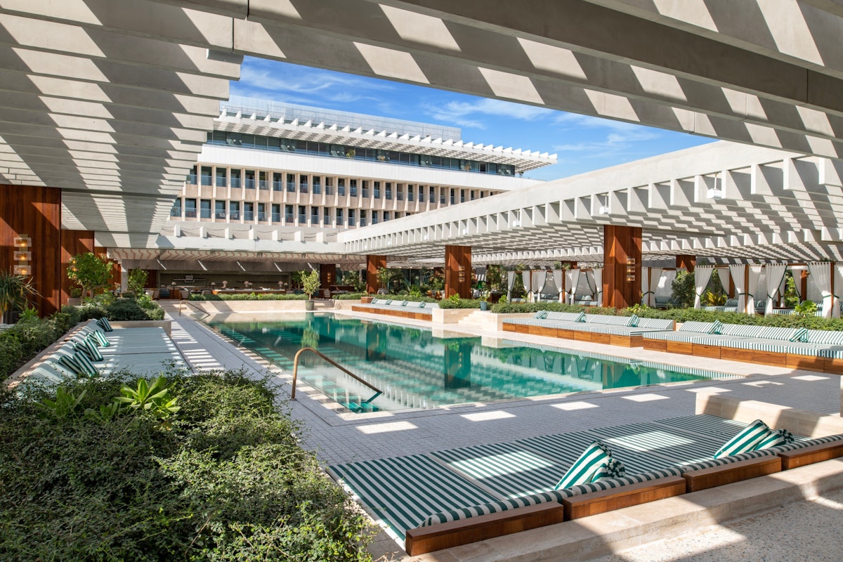 Modern pool area with lounge chairs and shaded pergolas, surrounded by lush greenery and a building.