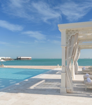 The Chedi Katara Hotel & Resort Beachfront pool with white cabanas, clear turquoise water, and a sunny sky.