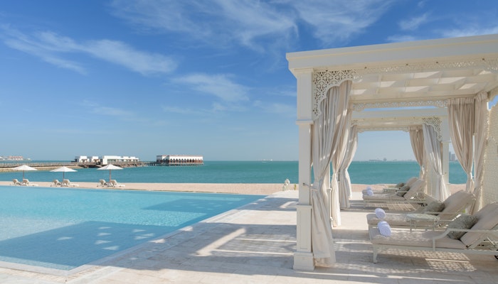 The Chedi Katara Hotel & Resort Beachfront pool with white cabanas, clear turquoise water, and a sunny sky.