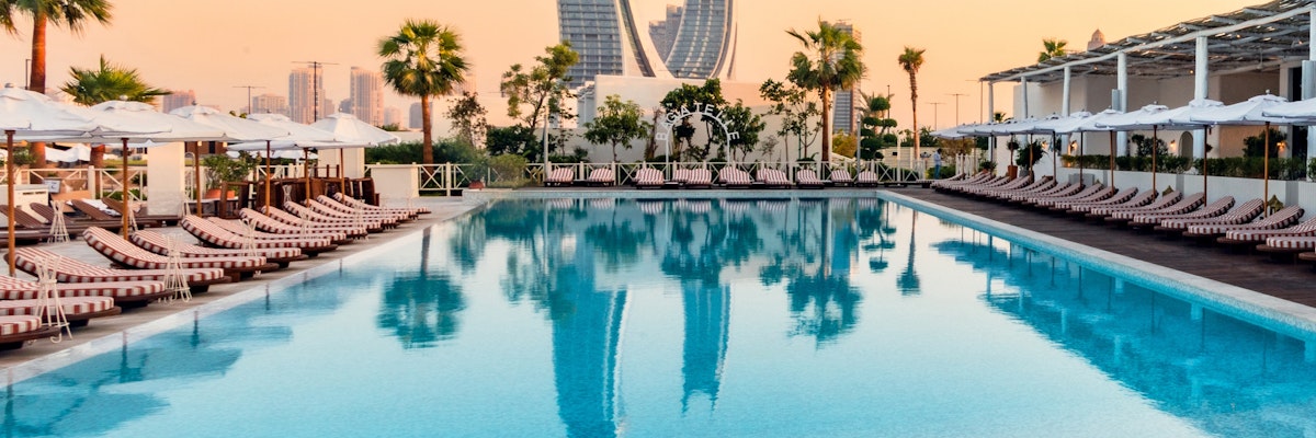 Bagatelle Beach Club Doha Luxury pool with lounge chairs, palm trees, and a city skyline with iconic skyscrapers at sunset.