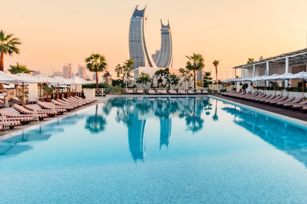 Bagatelle Beach Club Doha Luxury pool with lounge chairs, palm trees, and a city skyline with iconic skyscrapers at sunset.