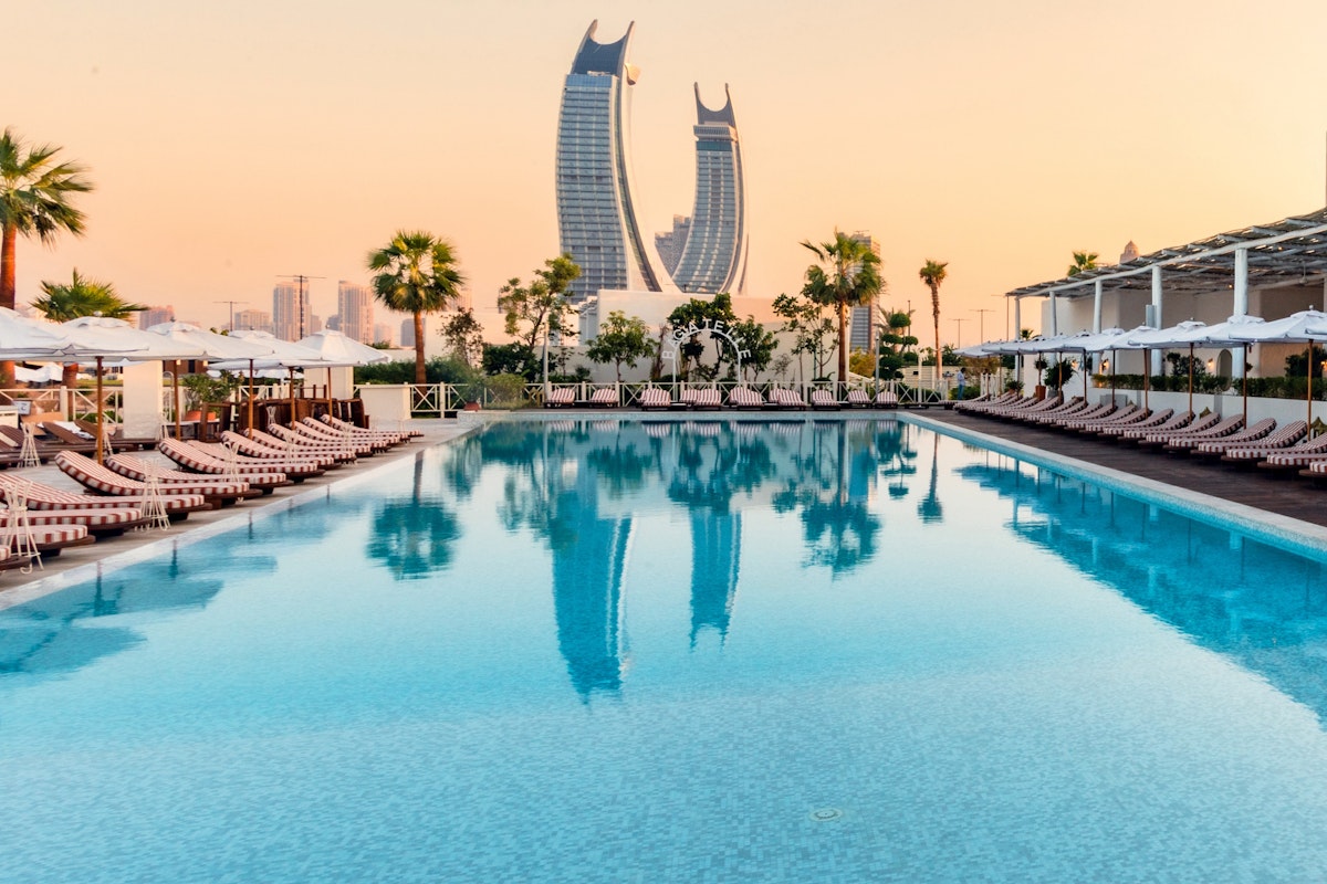 Bagatelle Beach Club Doha Luxury pool with lounge chairs, palm trees, and a city skyline with iconic skyscrapers at sunset.