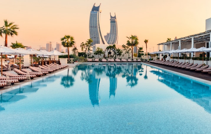 Luxury pool with lounge chairs, palm trees, and a city skyline with iconic skyscrapers at sunset.