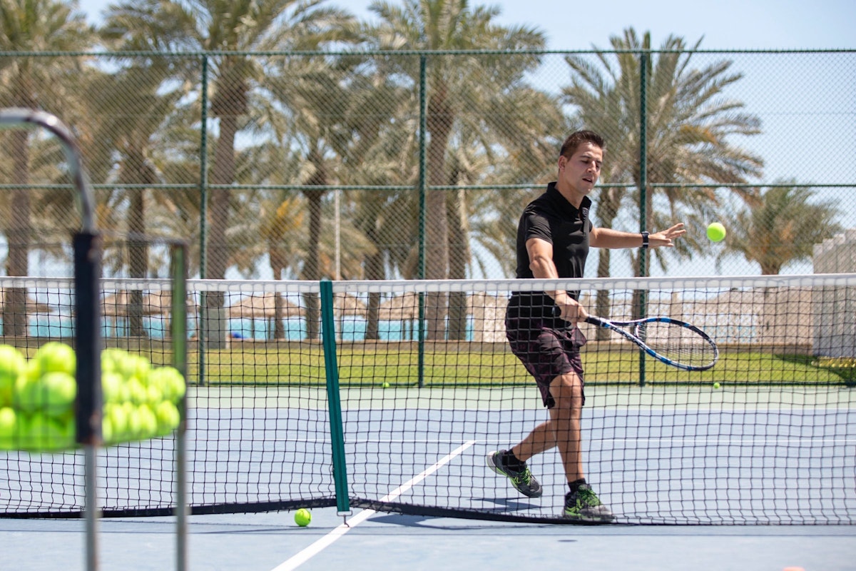 Tennis player hitting a ball on the court with a tennis racket, with palm trees in the background.