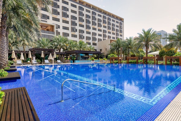 Outdoor pool with lounge chairs and umbrellas surrounded by palm trees and a hotel.
