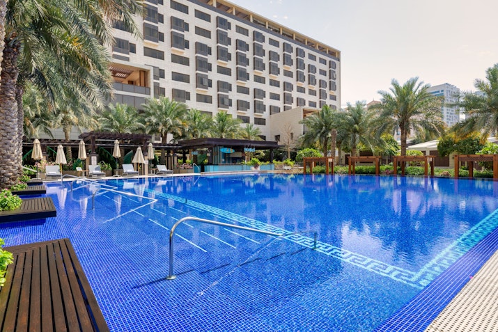 Outdoor pool with lounge chairs and umbrellas surrounded by palm trees and a hotel.