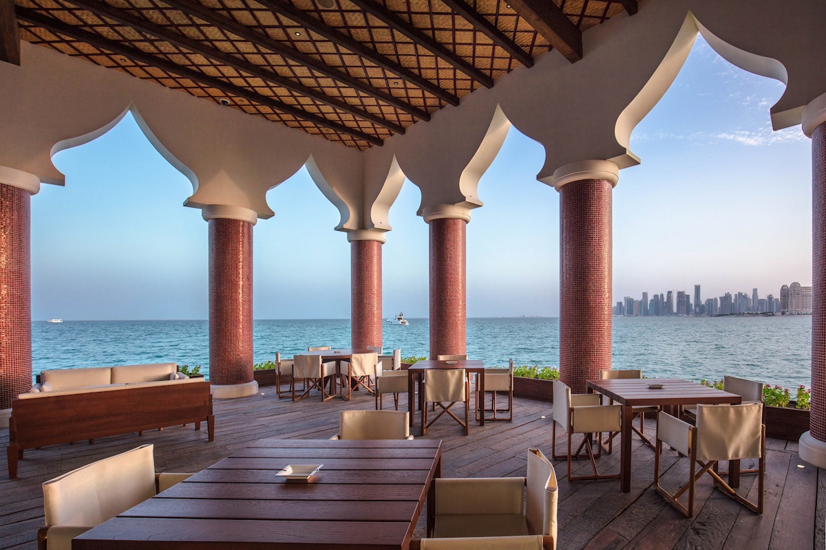 The Chedi Katara Hotel & Resort Outdoor dining area with wooden tables, overlooking the ocean and a city skyline in the distance.