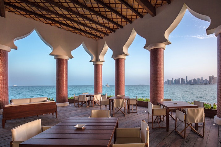 The Chedi Katara Hotel & Resort Outdoor dining area with wooden tables, overlooking the ocean and a city skyline in the distance.