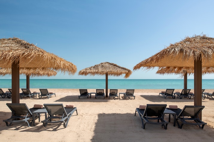 Beach with straw umbrellas, lounge chairs, and clear blue water under a sunny sky