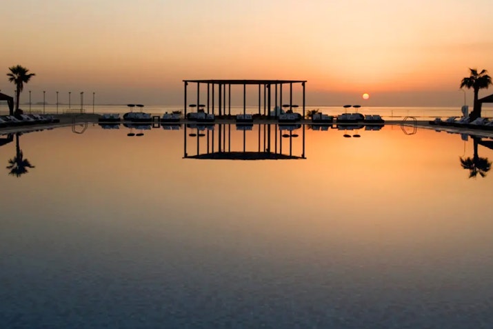 The St. Regis Doha Sunset reflecting on a tranquil pool, with a view of a modern gazebo structure and palm trees.