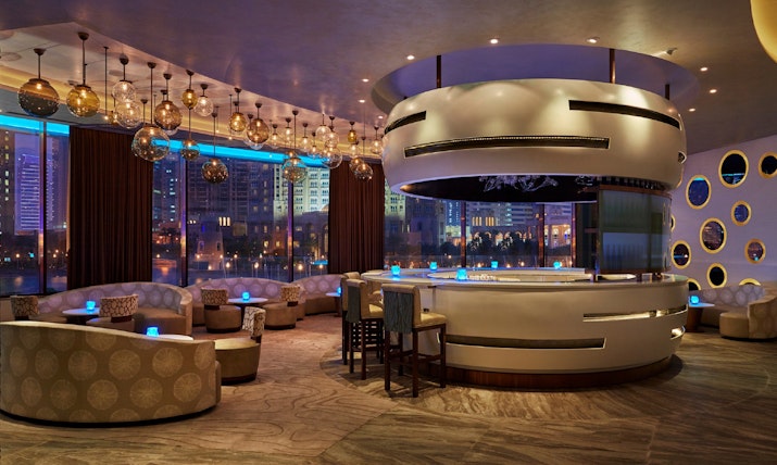 Sip and Savour Contemporary bar with circular seating, illuminated by pendant lights and panoramic city views, featuring a unique design.