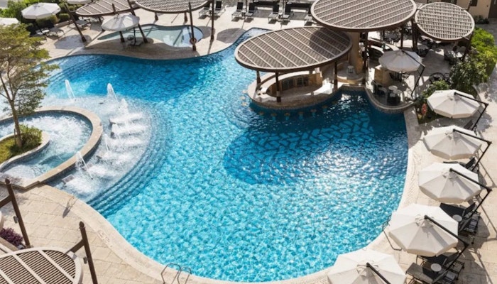 Hilton Doha The Pearl Swimming pool with circular cabanas, water fountains, and lounge chairs, surrounded by umbrellas and greenery.