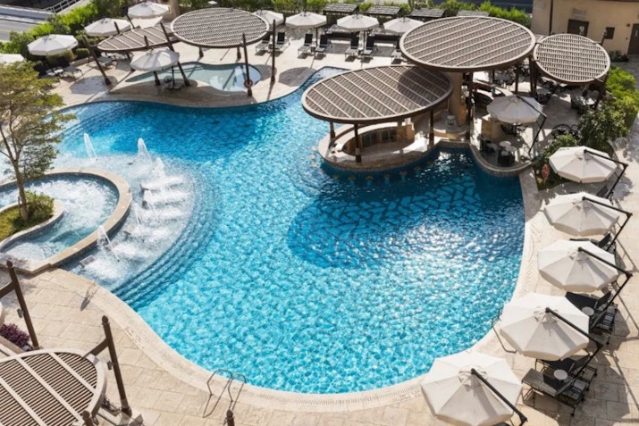 Swimming pool with circular cabanas, water fountains, and lounge chairs, surrounded by umbrellas and greenery.