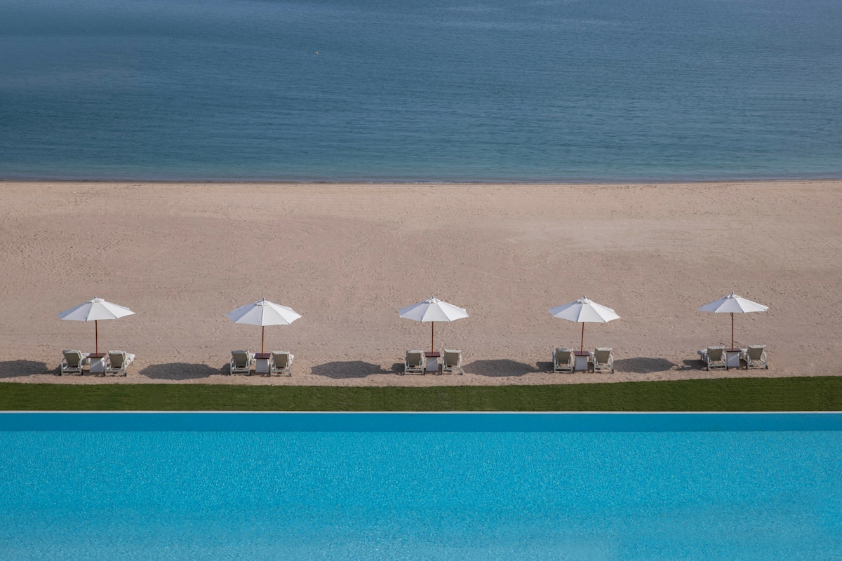 View of beach lounge chairs with umbrellas next to a swimming pool and ocean.