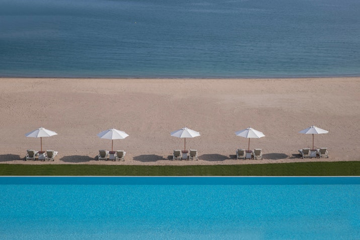 View of beach lounge chairs with umbrellas next to a swimming pool and ocean.