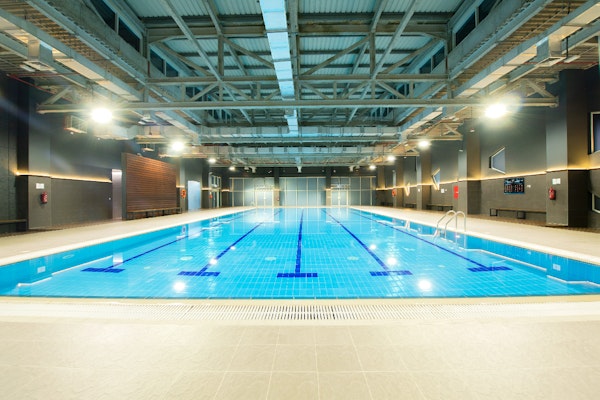 Indoor swimming pool with clear water and bright lighting, surrounded by a modern gym facility.
